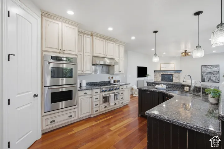 Kitchen featuring cream-colored cabinetry, stone countertops, stainless steel double wall ovens, a gas range with a stainless steel vent hood, and wood-finish flooring
