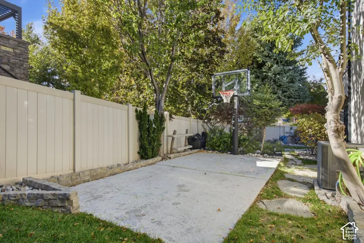 Concrete sports court featuring a mounted basketball hoop, enclosed by solid vinyl fencing and stone retaining walls, surrounded by mature landscaping