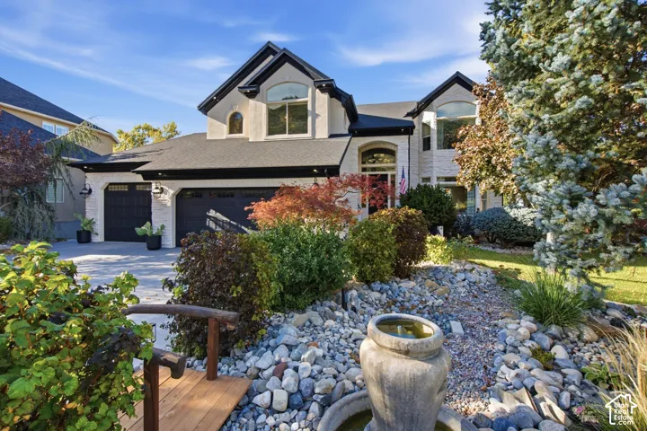 Multi-gabled exterior featuring light-toned masonry, a multi-car garage with dark-finish doors, arched window accents, and front-yard landscaping including a stone-lined garden with a decorative fountain and timber pedestrian bridge