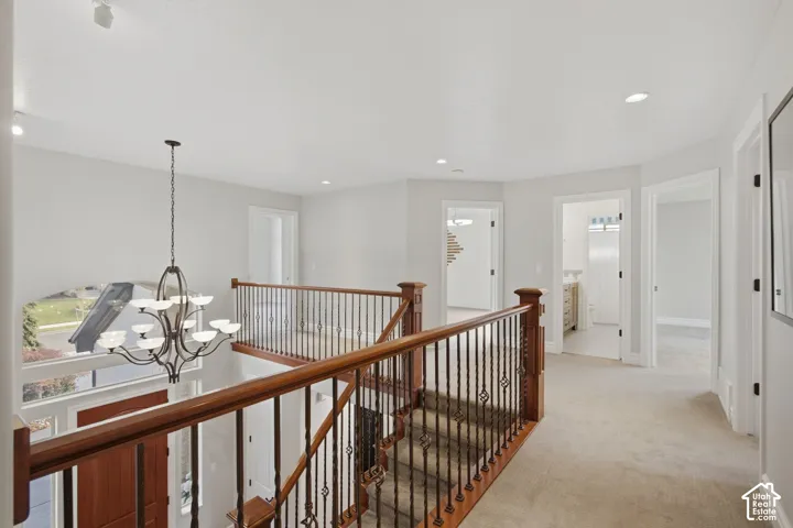 Open hallway featuring neutral carpeting, a staircase with a wooden handrail and decorative metal balusters, recessed lighting, and a multi-arm chandelier