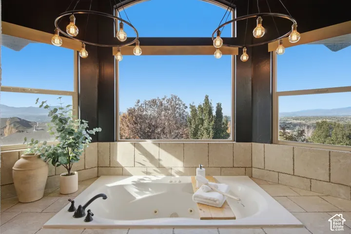 Jetted soaking tub with matte black faucet, surround of neutral square tile, oversized windows, and dual circular pendant light fixtures