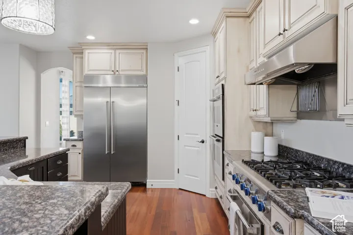 Kitchen featuring wood-finish flooring, stone countertops, stainless steel appliances including a double wall oven and gas range, custom cabinetry, and recessed lighting