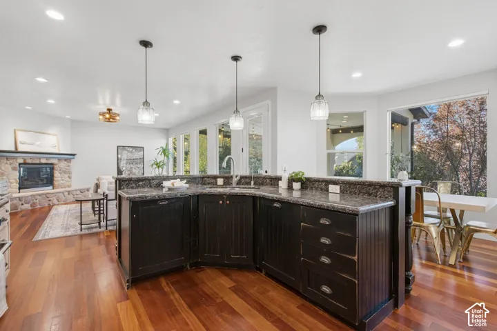 Kitchen featuring dark cabinetry with beadboard detailing, stone-surface countertops, wood-finish flooring, three glass pendant lights, and a stone-clad fireplace