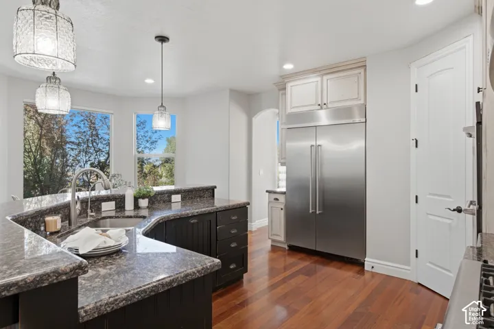 Kitchen featuring dark-toned cabinetry, stone-finish countertops, stainless steel built-in refrigerator, wood-finish flooring, and multi-light pendant fixtures