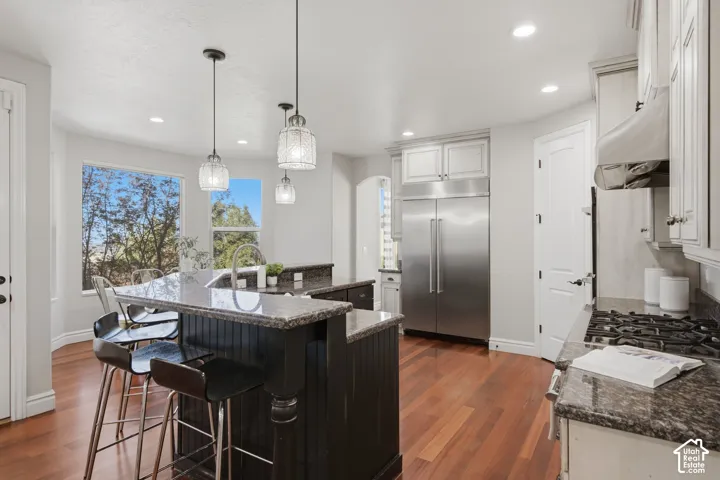 Kitchen featuring wood-finish flooring, a raised breakfast bar with stone surfacing, stainless steel double-door refrigerator, gas cooktop with an integrated range hood, and pendant lighting
