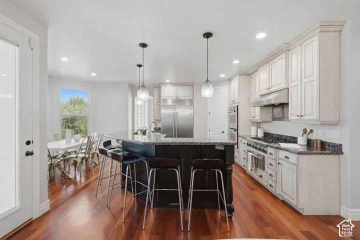 Kitchen featuring wood-finish flooring, a central island with a contrasting dark base, white cabinetry, stainless steel built-in appliances, and recessed and pendant lighting