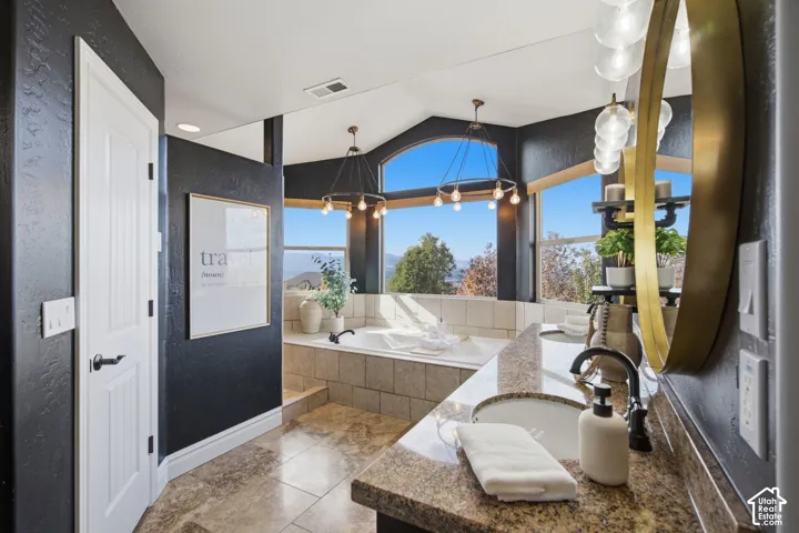Bathroom featuring a stone-topped dual vanity with undermount sinks, tile-surround soaking tub, arched window, and custom light fixtures