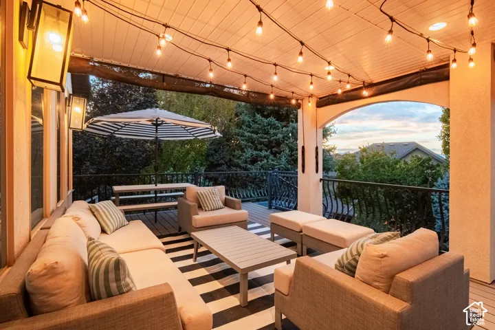 Covered outdoor deck with string lighting, exterior wall sconces, wood-finish decking, and wrought iron railing