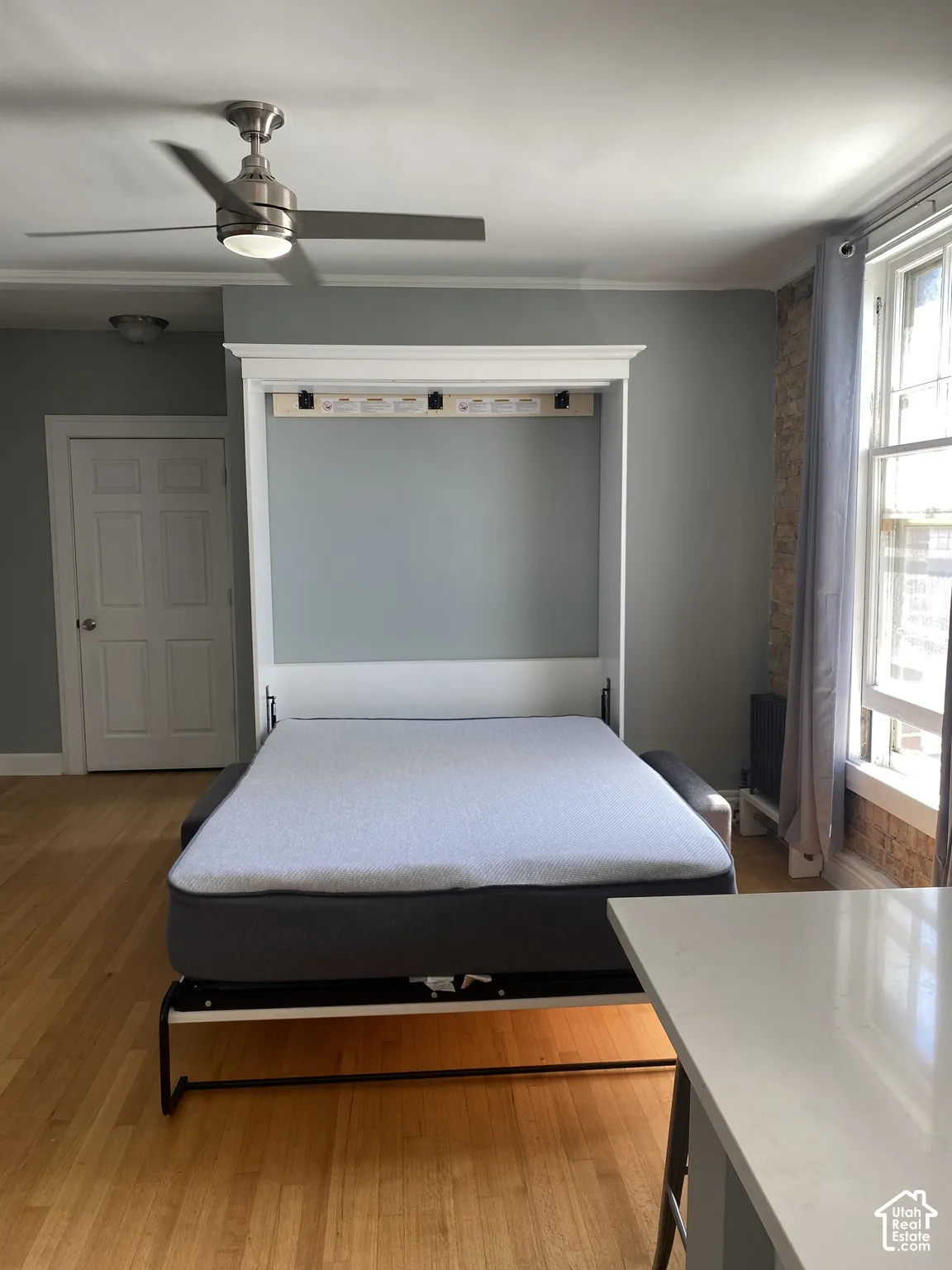 Wood-finish flooring, built-in Murphy bed, satin nickel ceiling fan with integrated lighting, exposed brick wall detail, and a large window with white trim