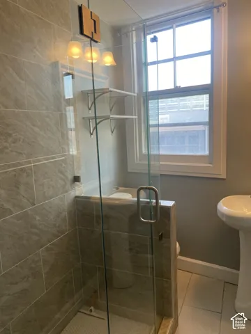 Tiled shower with a glass enclosure, pedestal sink, wall-mounted light fixture, and window with a window treatment rod