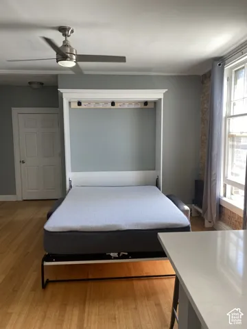Wood-finish flooring, built-in Murphy bed, satin nickel ceiling fan with integrated lighting, exposed brick wall detail, and a large window with white trim