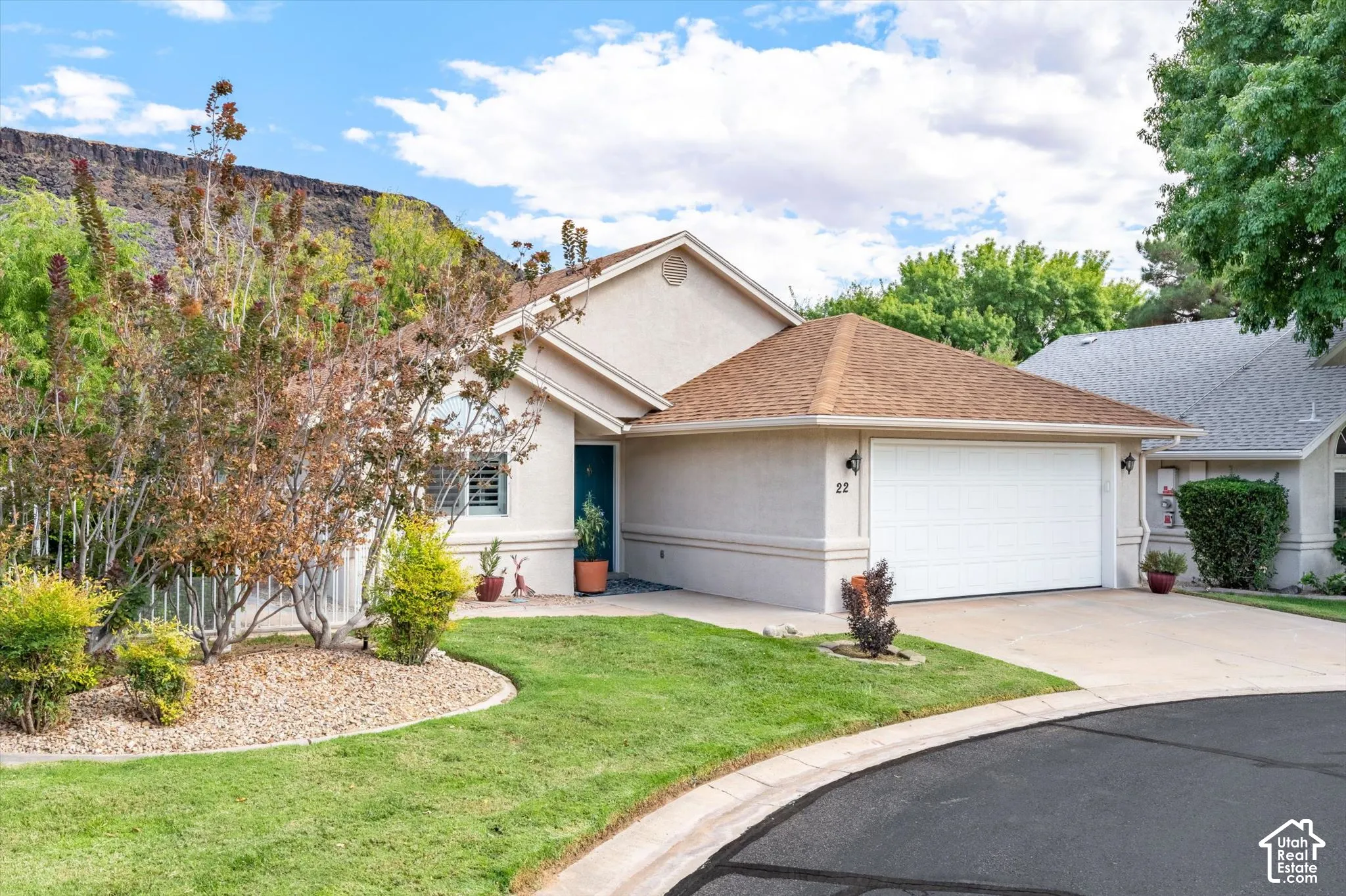 Stucco exterior, attached two-car garage, pitched shingle roof, landscaped front yard with lawn and rock features, and mature trees