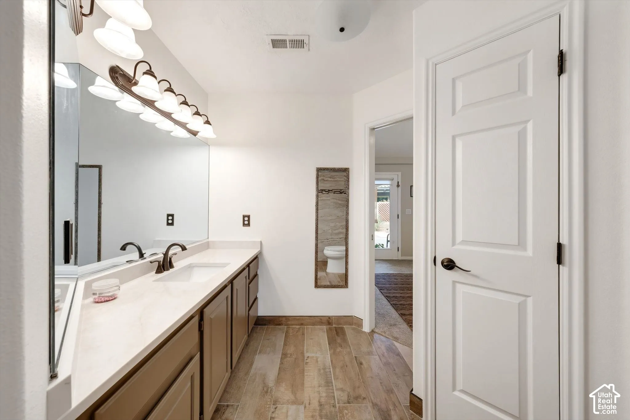Extended vanity with dual sinks, overhead lighting, wood-finish flooring, and a full-length mirror