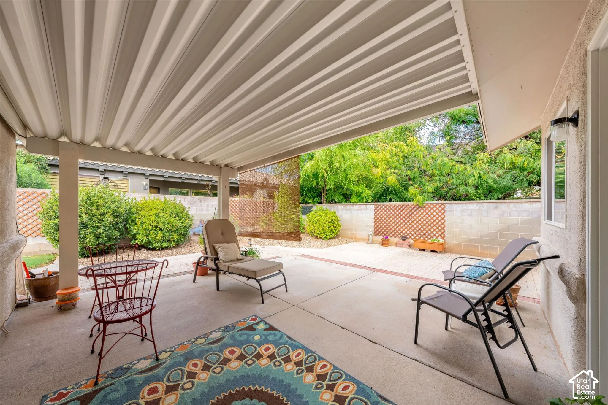Expansive covered patio featuring a corrugated overhead structure, concrete flooring with brick accents, and a privacy lattice