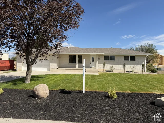 Single-story residence featuring light-colored brick exterior, attached garage, and a front lawn with mature trees and landscaped beds