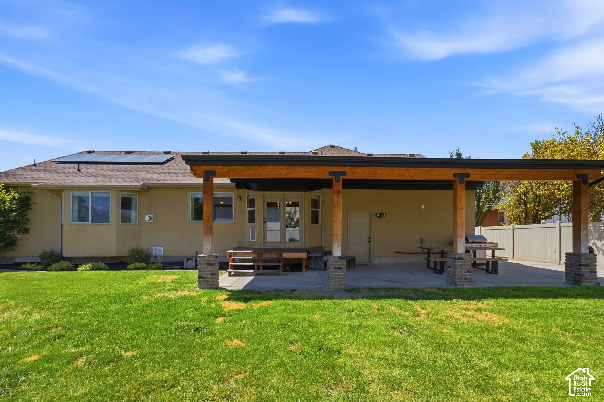 Expansive rear exterior featuring a stucco facade, a covered patio with wood beams and stone-clad columns, a concrete patio surface, a manicured lawn, and solar panels on the roof