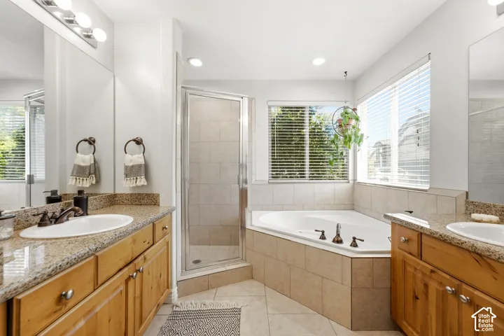 Bathroom featuring dual vanities with granite-style countertops, wood cabinetry, a tiled corner soaking tub, a separate glass-enclosed shower, and windows with horizontal blinds