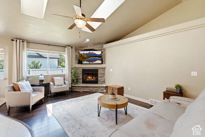 Living room featuring a vaulted ceiling with skylights, wood-finish flooring, a stacked stone fireplace, and a ceiling fan