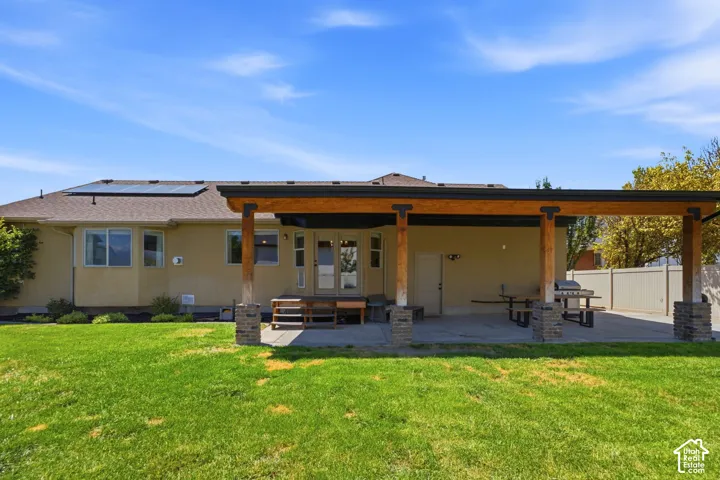 Expansive rear exterior featuring a stucco facade, a covered patio with wood beams and stone-clad columns, a concrete patio surface, a manicured lawn, and solar panels on the roof