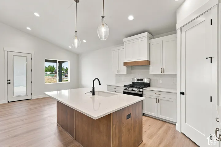 Kitchen featuring a center island with a built-in sink and black faucet, stainless steel gas range, white cabinetry, and wood-finish flooring