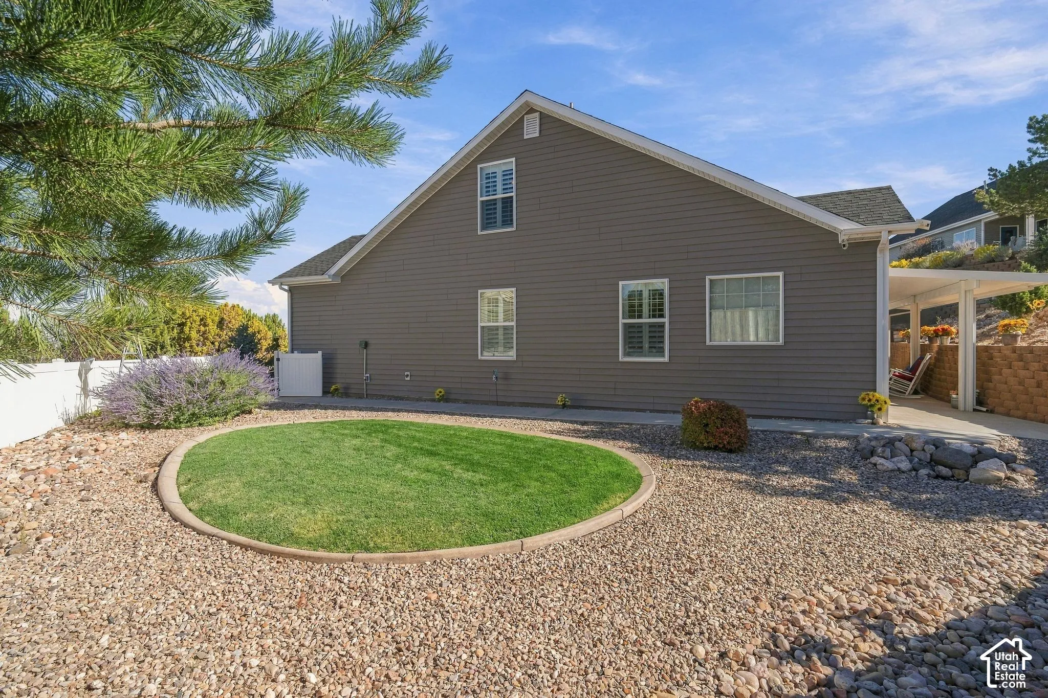Exterior siding, multiple windows with white frames, covered patio structure, gravel landscaping, and a circular lawn feature