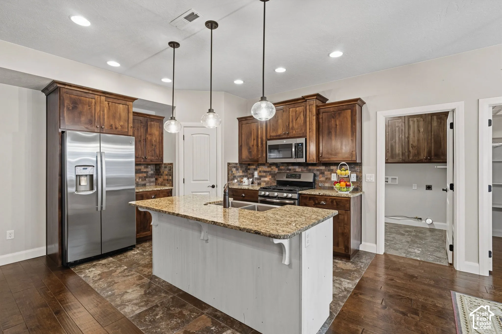 Kitchen featuring wood cabinetry, stainless steel appliances, a center island with a built-in sink, and pendant lighting