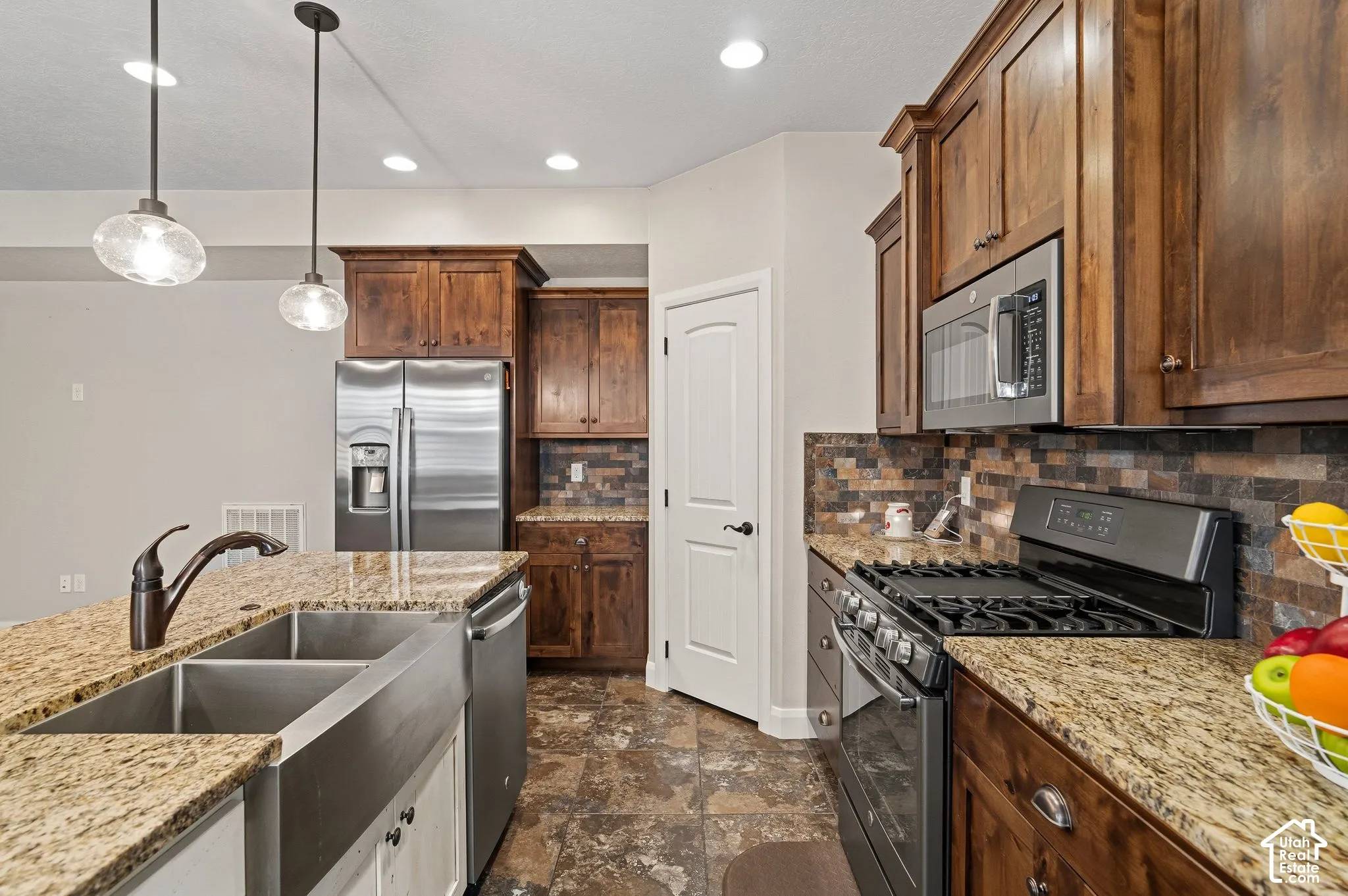 Galley-style kitchen featuring rich wood cabinetry, granite countertops, stainless steel appliances including a gas cooktop and built-in microwave, undermount sink with chrome faucet, and brick tile backsplash