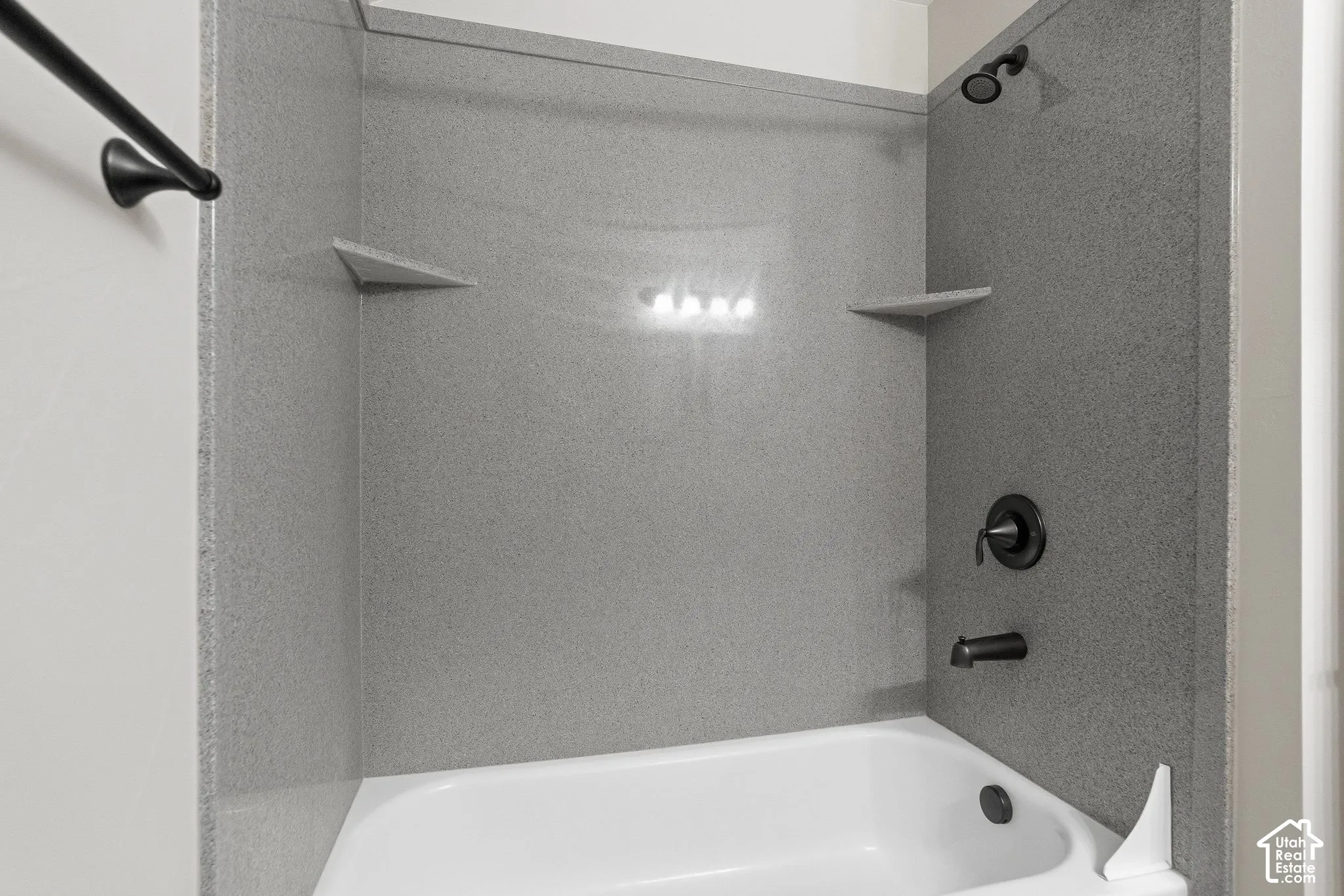 Bathtub with textured wall surround, matte black shower fixtures, and built-in corner shelving