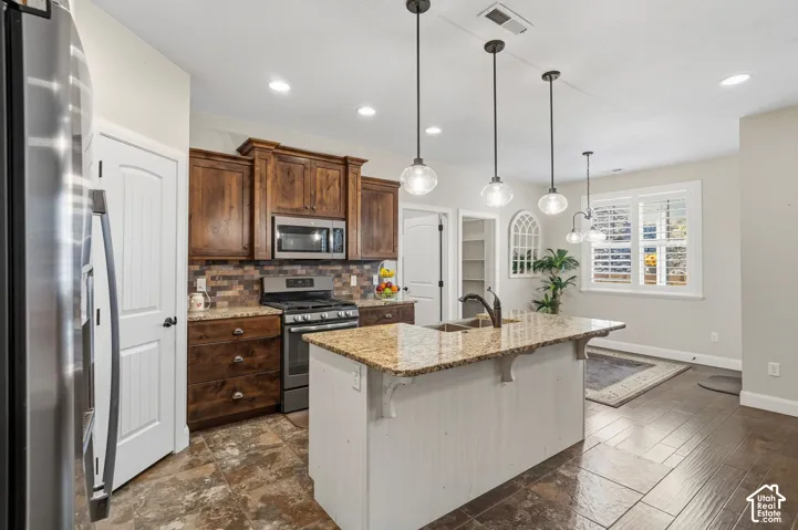 Kitchen featuring wood-finish cabinetry, stainless steel appliances, granite countertops, an island with a built-in sink, and pendant lighting