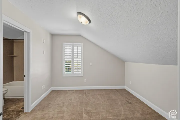 Sloped ceiling room featuring carpet flooring, natural light from a window with plantation shutters, and a flush mount ceiling light fixture