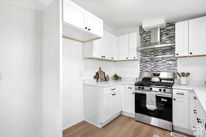 White cabinetry with black hardware, white countertops, wood-finish flooring, stainless steel range with overhead hood, and a mosaic tile backsplash