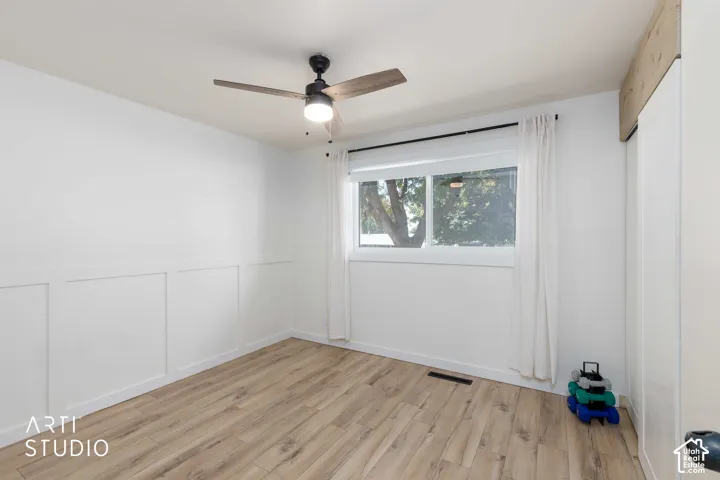 Wood-finish flooring, white panel wall detailing, window with white trim, and a ceiling fan with light fixture