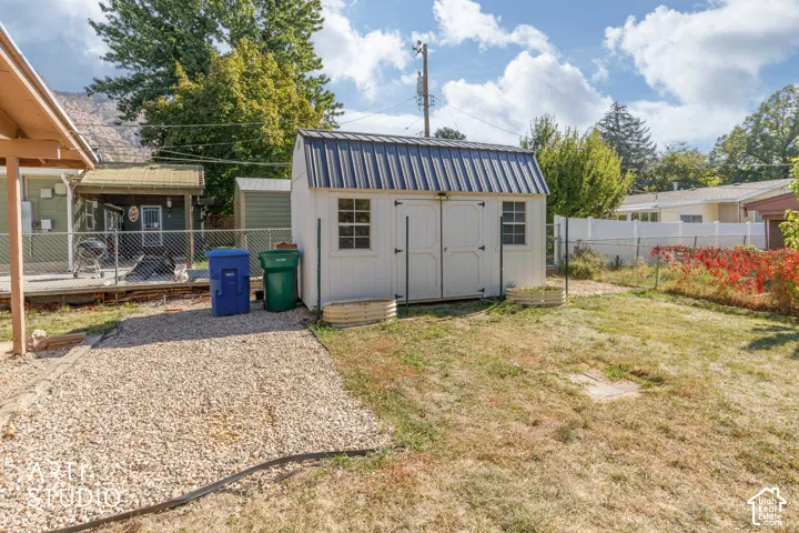 White storage shed with a dark metal roof, featuring a gravel pathway and a partial chain-link fence