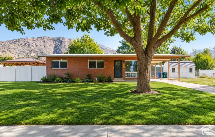 Single-level brick exterior featuring a covered carport, mature landscaping, white vinyl fencing, and a detached storage structure set against a mountain backdrop