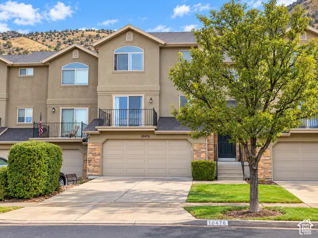 Three-story stucco exterior with stone accents, featuring an integrated two-car garage, a second-story balcony with iron railing, and a mature tree in the front yard