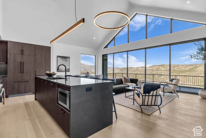 Open-concept living space with vaulted ceilings, expansive floor-to-ceiling windows, light wood-finish flooring, a contemporary kitchen island with an integrated microwave, and built-in dark wood-finish cabinetry