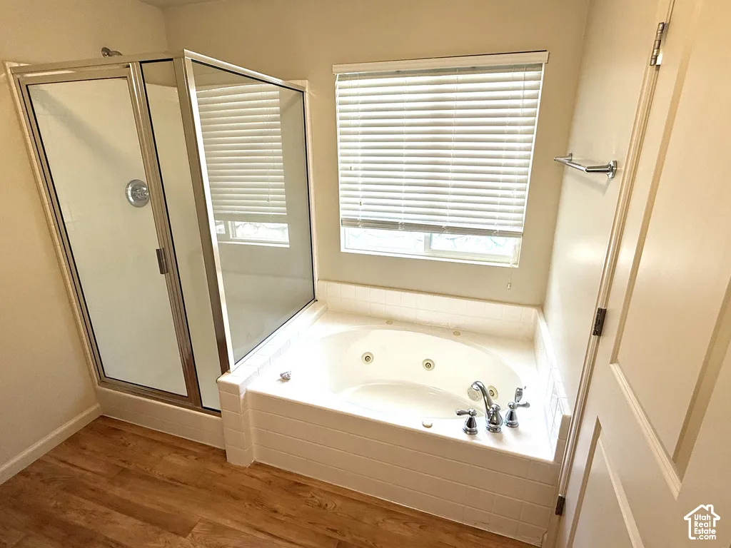 Jetted soaking tub, separate shower enclosure with glass doors, wood-finish flooring, and window with blinds