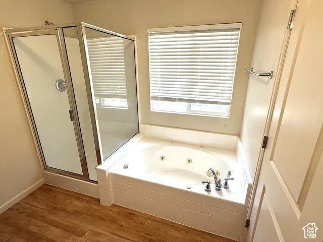 Jetted soaking tub, separate shower enclosure with glass doors, wood-finish flooring, and window with blinds