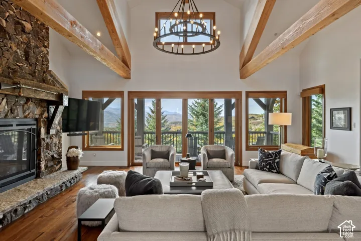 Vaulted ceilings with exposed wood beams, a stone fireplace, wood-finish flooring, a large circular chandelier, and extensive windows with wood trim