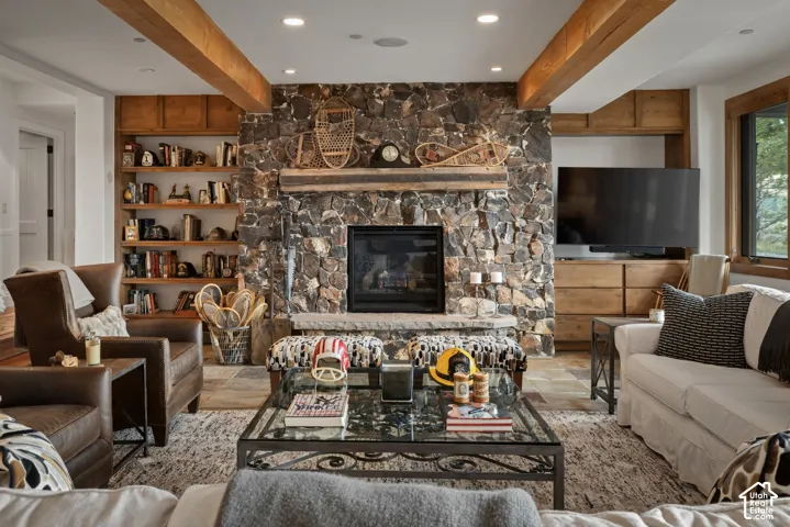 Exposed wood beams, stone fireplace, built-in shelving, wood-framed window, and recessed lighting