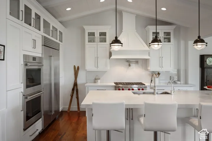 White cabinetry with glass-front uppers, stainless steel double ovens, large kitchen island with integrated sink, vaulted ceiling with exposed wood-finish paneling, and pendant lighting