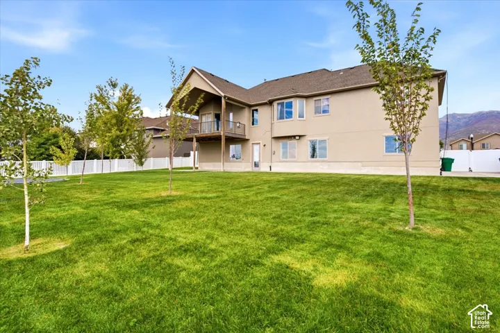 Expansive green lawn, stucco exterior, upper-level balcony, white vinyl fencing, and multiple windows