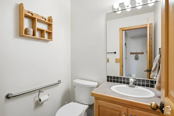 Vanity with wood-finish cabinetry and integrated sink, black tile backsplash, and a wall-mounted mirror with overhead light fixture