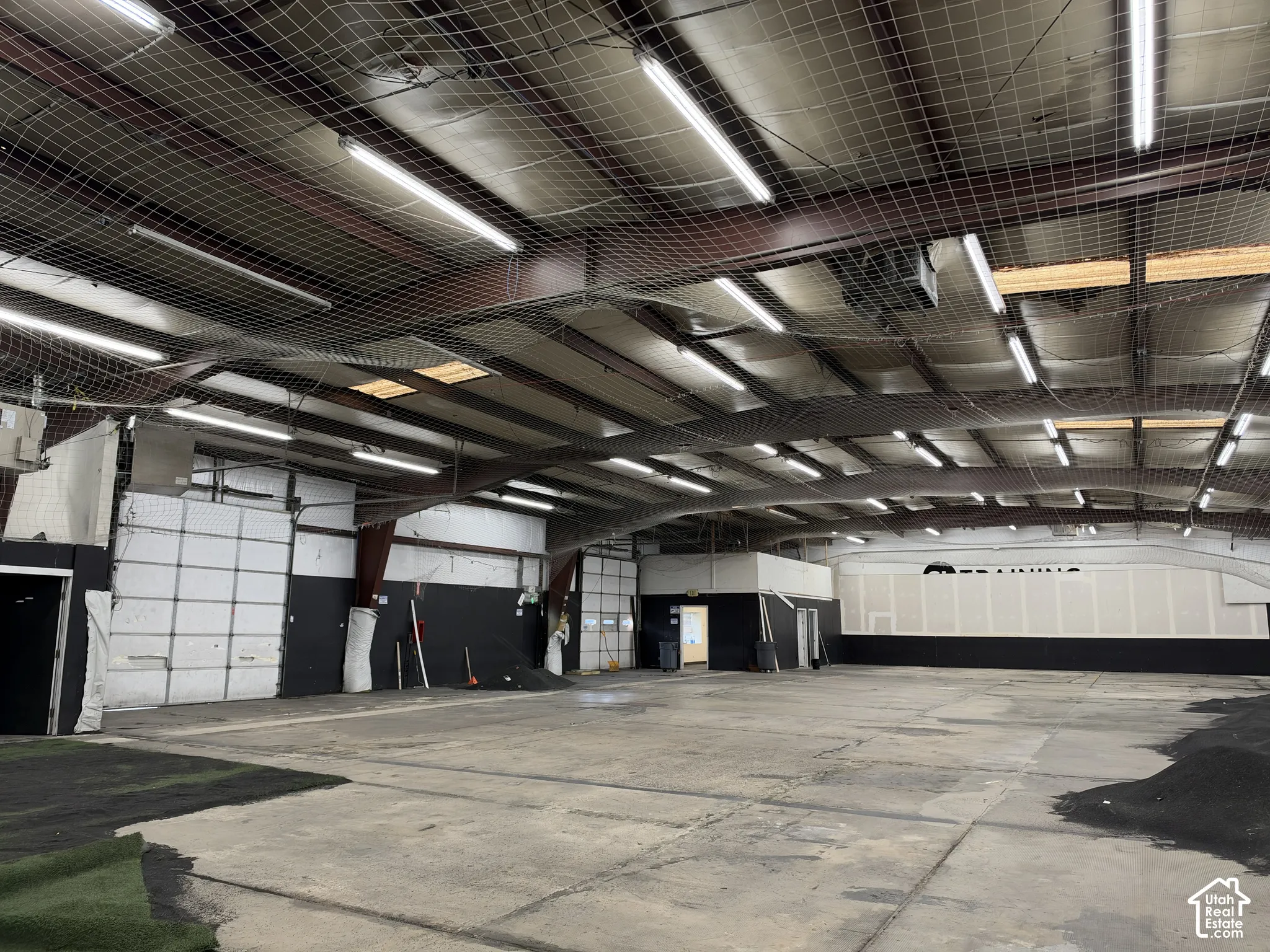 Expansive interior space featuring exposed structural beams, fluorescent lighting, high ceilings with protective netting, and multiple roll-up doors