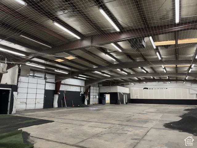 Expansive interior space featuring exposed structural beams, fluorescent lighting, high ceilings with protective netting, and multiple roll-up doors