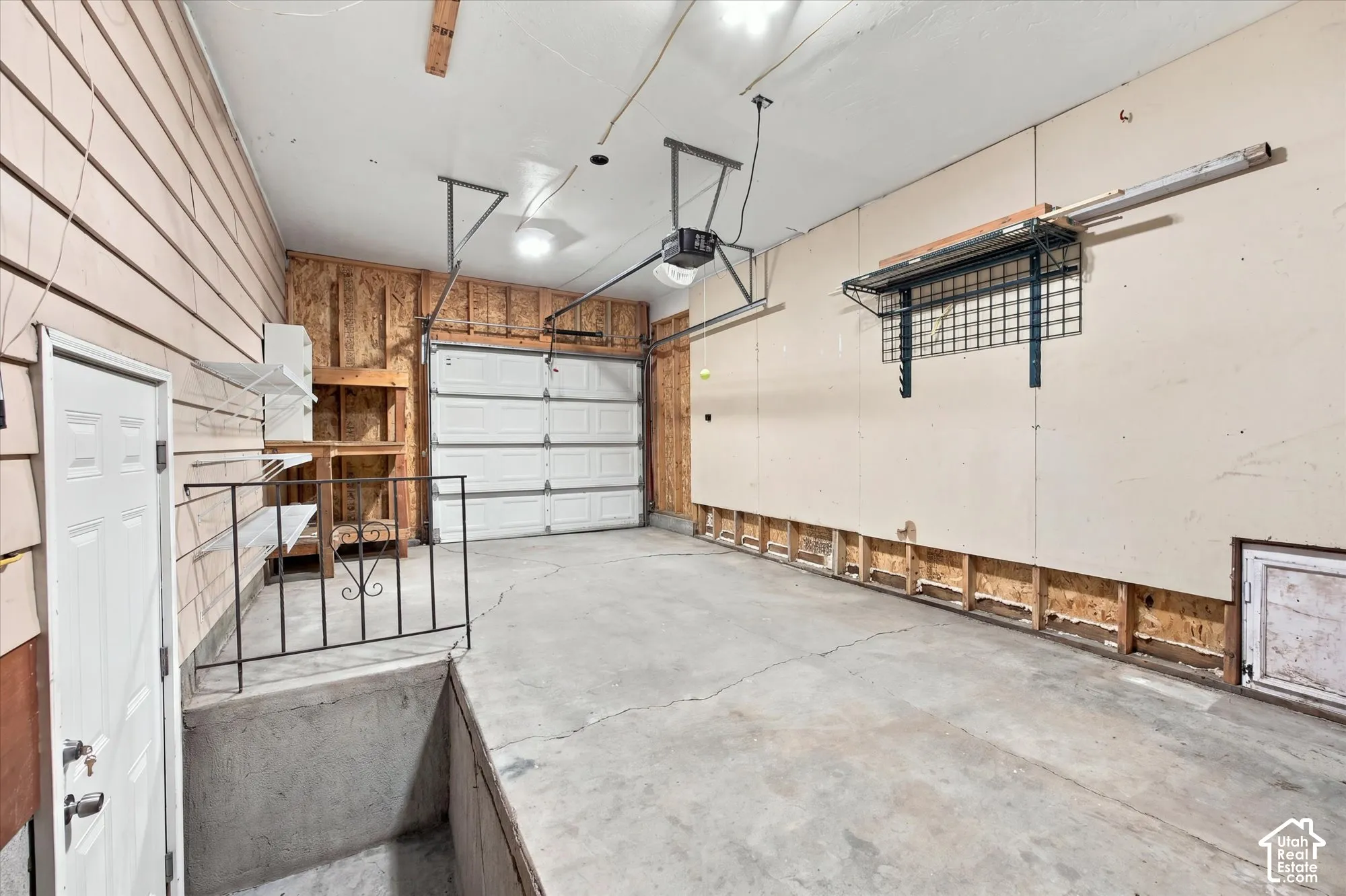 Concrete garage flooring, single garage door, overhead lighting, built-in shelving, wall-mounted storage rack. Basement entrance from Garage