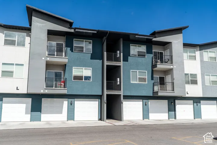 Modern multi-unit residential building featuring clean architectural lines, mixed exterior finishes in blue-grey and white tones, expansive window placement throughout, individual garage spaces on the ground level, and contemporary balconies with metal ra