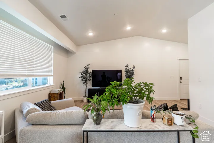 Modern living space featuring recessed ceiling lighting, large window with motorized roller shades, wall-mounted flat-screen television, light wood-finish flooring, and fresh indoor greenery throughout the room