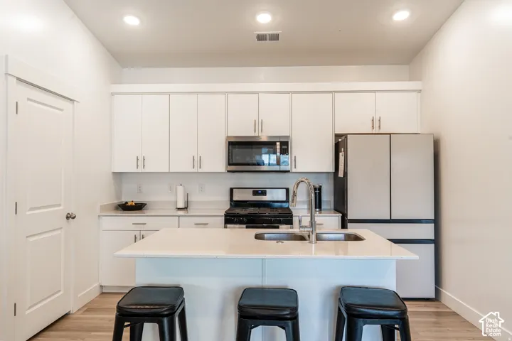 Kitchen featuring white cabinetry, stainless steel appliances, a center island with sink, light-toned countertops, and wood-finish flooring