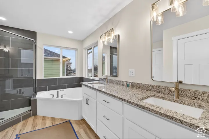 Dual sink vanity with granite countertop, wood-finish flooring, a soaking tub, and an enclosed shower with dark subway tile